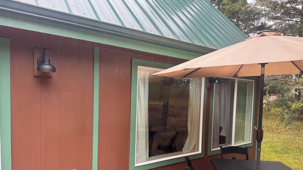 A wooden deck with a table and chairs, a window with curtains, and a green metal roof.