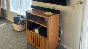 A room with a flat-screen TV mounted on the wall, a wooden entertainment center with a shelf, a window with white curtains, and a small basket on the floor.