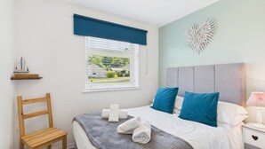A bedroom with a bed, a chair, a window with blinds, a decorative wall piece, and a nightstand with a lamp. - Bayview 62 - 2 Bedroom Chalet - Bayview Oxwich (Swansea)