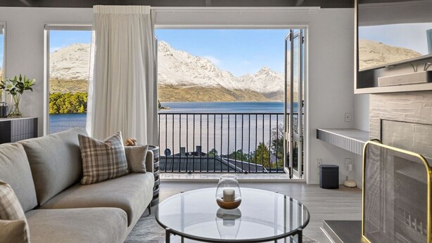 Una sala de estar moderna con un sofá, una mesa de centro de vidrio y una televisión. Ventanas amplias ofrecen vistas a un lago y montañas.
