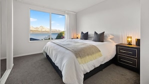 Un dormitorio con una cama grande, una mesita de noche, un ventanal con vistas a montañas y al agua, y piso alfombrado.