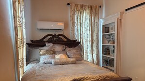 Un lit bien fait, avec une tête de lit, un climatiseur fixé au mur et une étagère ornée d’objets décoratifs.