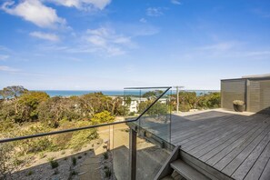 A wooden deck with a glass balustrade overlooking a coastal landscape with trees and buildings. - Stunning Roadknight Residence (Anglesea)