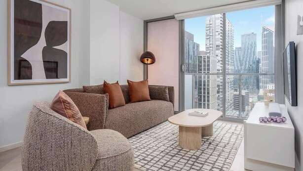 Una sala de estar moderna con sofá, sillón, mesa de centro y vista a la ciudad a través de amplios ventanales.