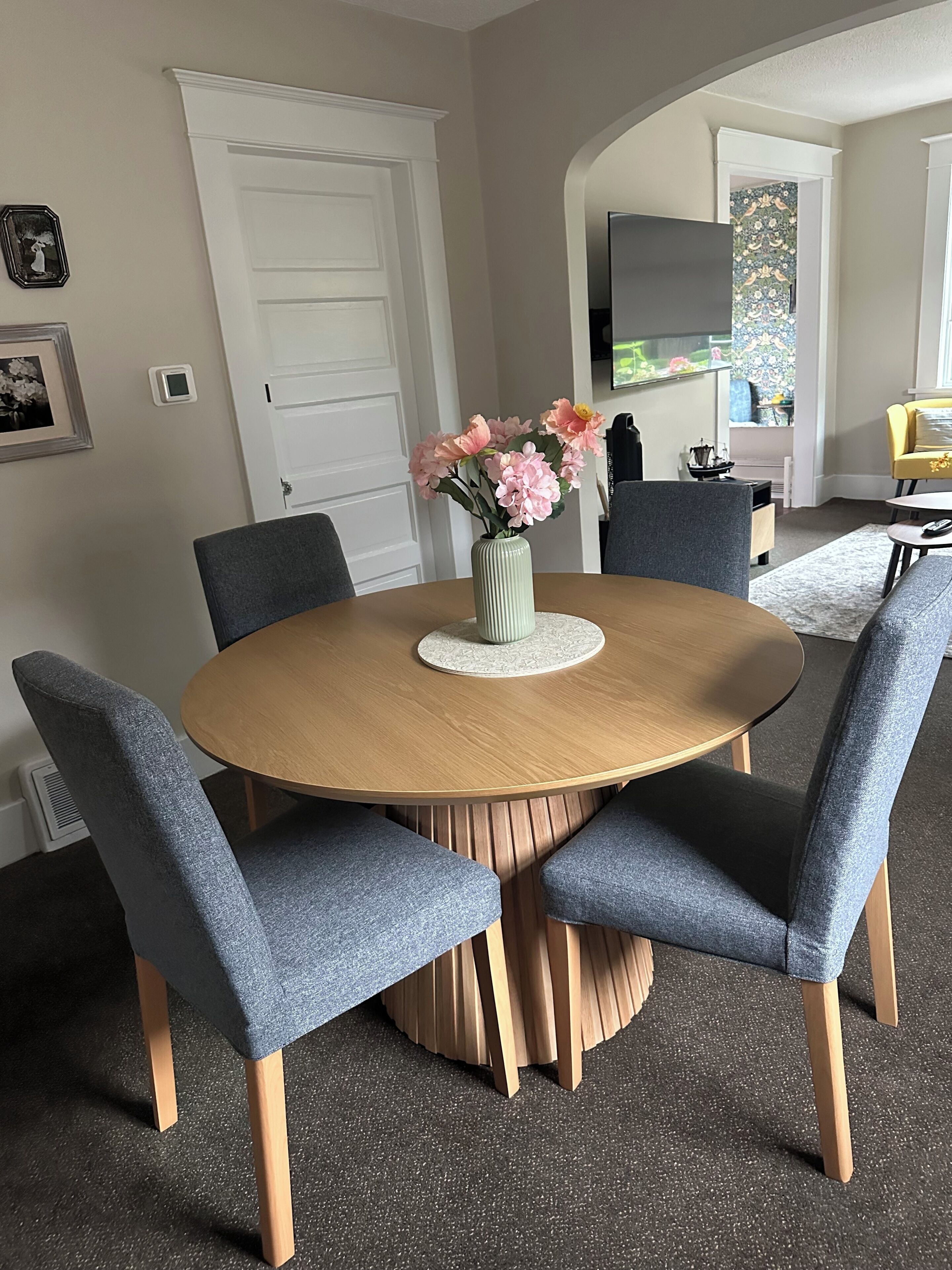 A round dining table with six grey upholstered chairs, a vase with pink flowers, and a white door in the background.