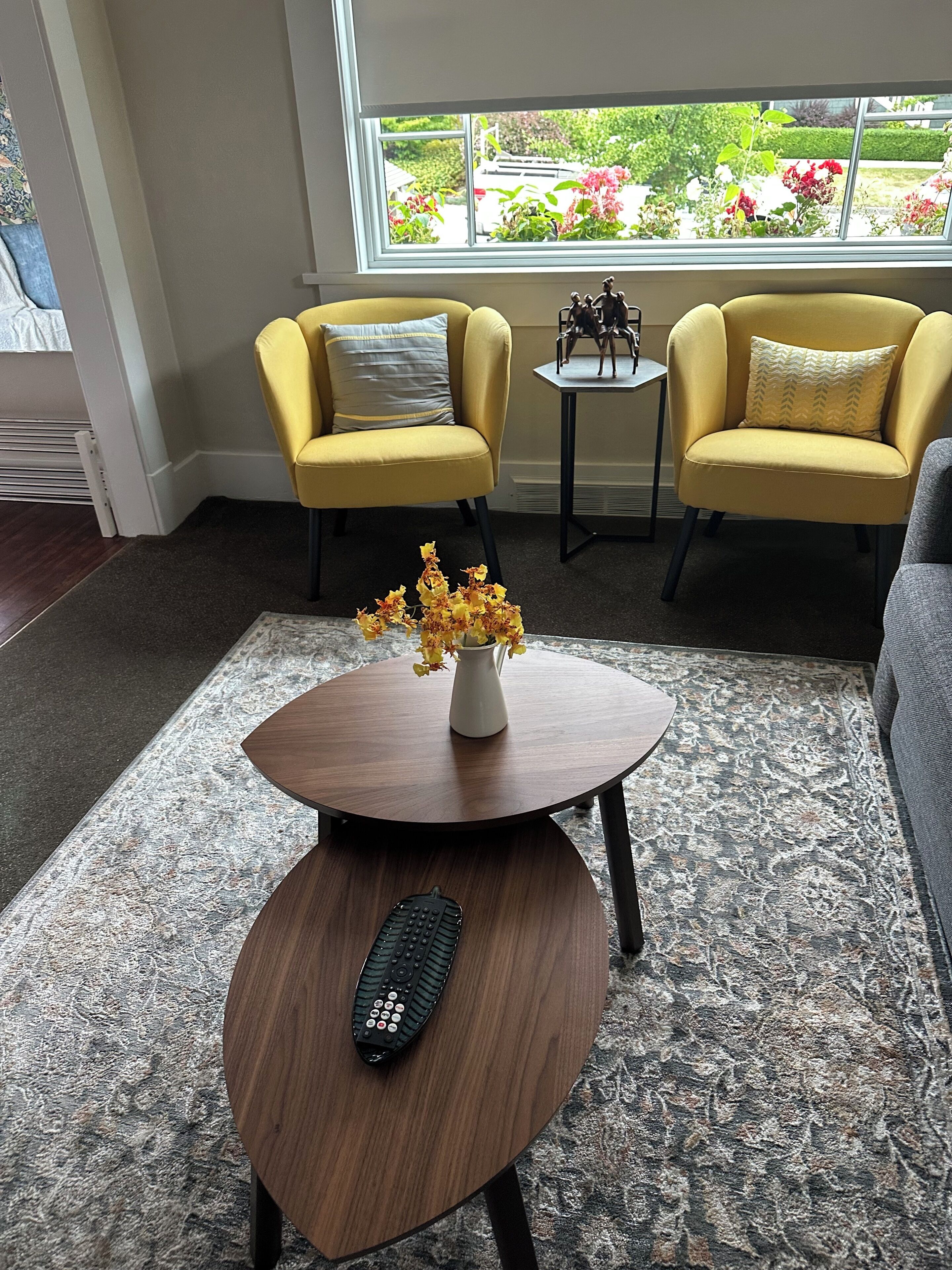 A living room with two yellow armchairs, a coffee table with a vase of flowers, a remote control, and a view of a garden through a window.