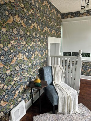 A room with a patterned wallpaper, a white chair, a small table with a lamp, and a radiator.