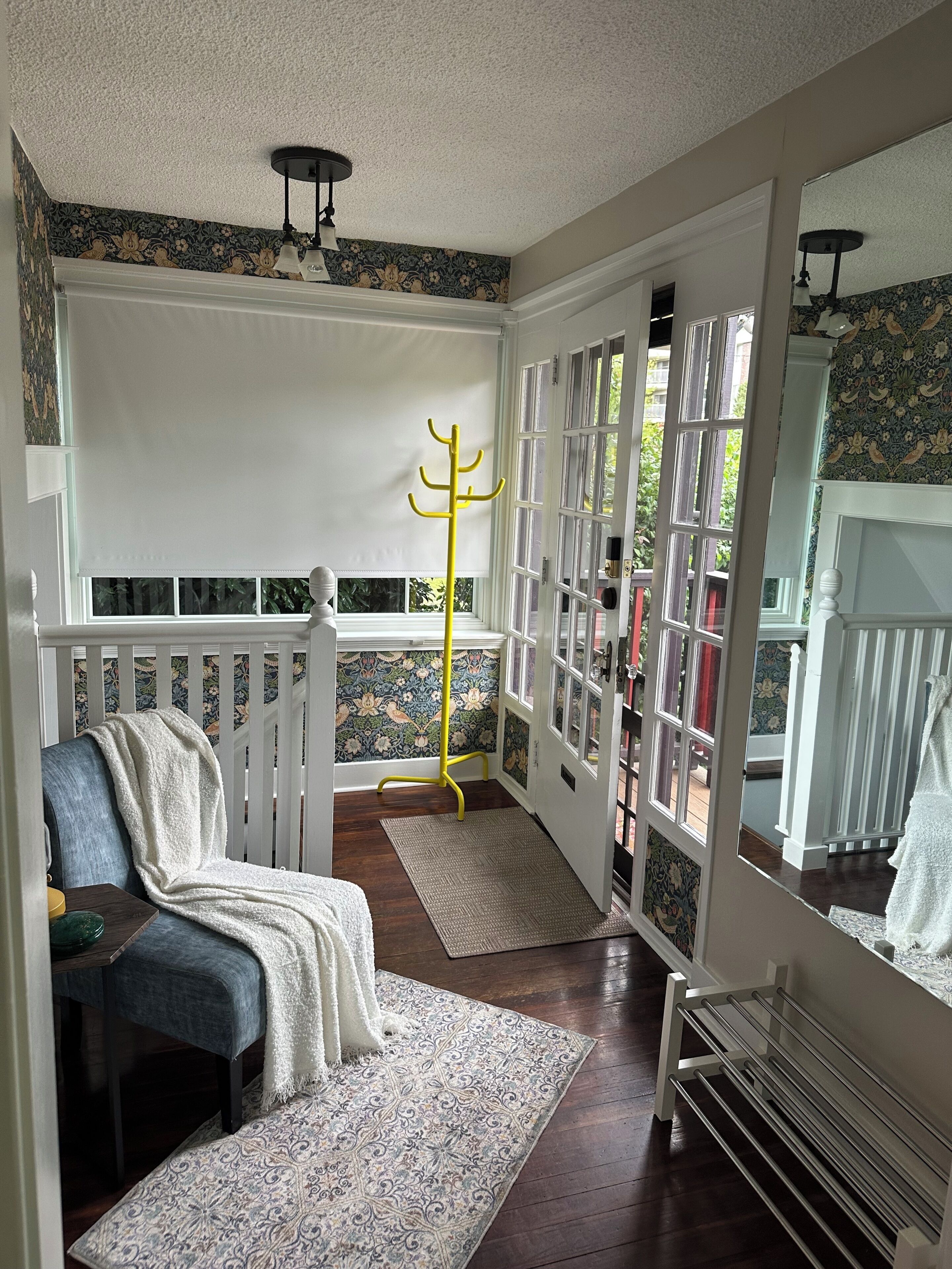 A room with a white chair, a yellow coat rack, a patterned rug, and a view through a glass door.