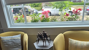 Dos sillones amarillos con cojines estampados, una mesita con una figurita, y una ventana con vista a coches aparcados y follaje.
