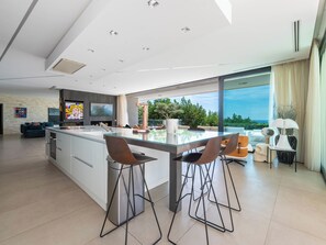A modern kitchen with a large island, bar stools, and a dining area with a view. - Contemporary Villa – Panoramic Sea View & Rooftop Jacuzzi (Mandelieu-la-Napoule)