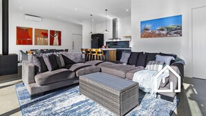 Een moderne woonkamer met een hoekbank, een eethoek met stoelen, een keuken en een groot schilderij aan de muur.