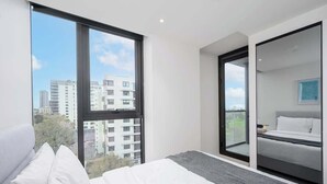 Una habitación de hotel moderna con un ventanal grande, una cama con ropa de cama blanca, una alfombra gris y vistas a edificios de la ciudad.