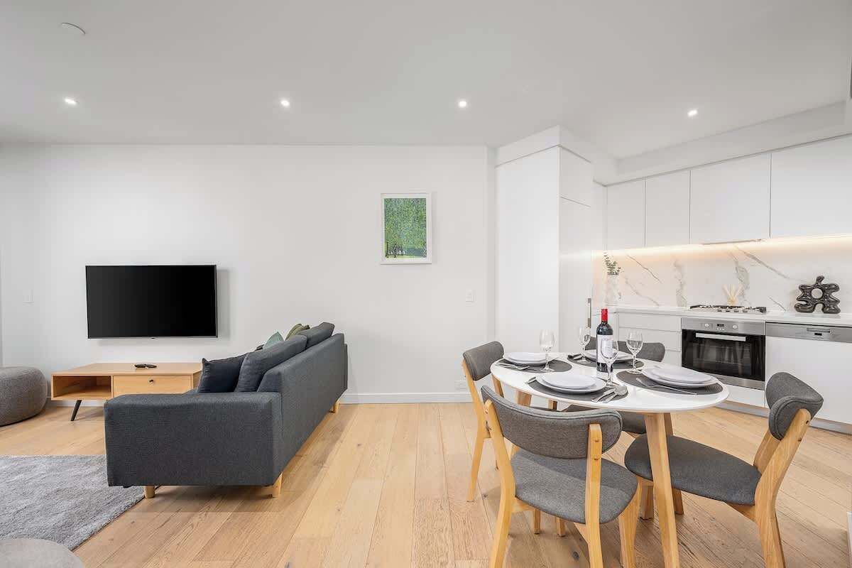 Una sala de estar moderna con televisor de pantalla plana, sofá gris, mesa de comedor con sillas y una zona de cocina con gabinetes blancos y encimera de mármol.