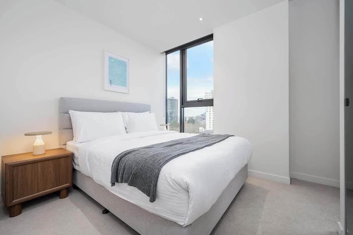 A modern bedroom with a large bed, a bedside table with a lamp, a window with a city view, and a framed picture on the wall.