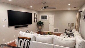 A living room with a flat-screen TV, a sofa, a dining table with chairs, a ceiling fan, and a window with blinds.