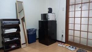 Una habitación con piso de madera, un refrigerador negro, un microondas, un cesto de basura azul, un estante de madera con objetos doblados y un par de pantuflas.