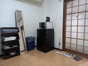 Una habitación con piso de madera, un refrigerador negro, un microondas, un cesto de basura azul, un estante de madera con objetos doblados y un par de pantuflas.