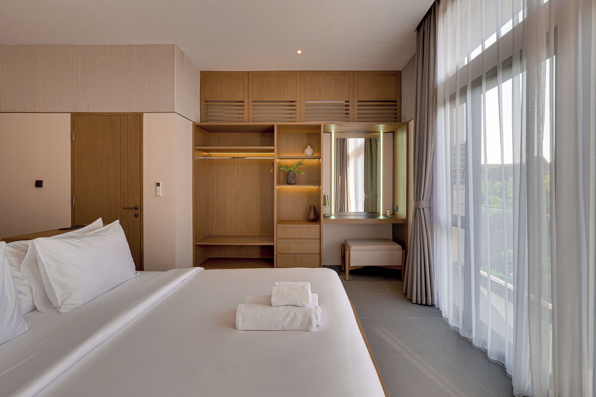 A hotel room with a large bed, a walk-in closet, a seating area, and a view through sheer curtains.