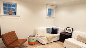 Een woonkamer met een bruine fauteuil, een witte bank, een beige vloerkleed en een bijzettafeltje met een fotolijstje.