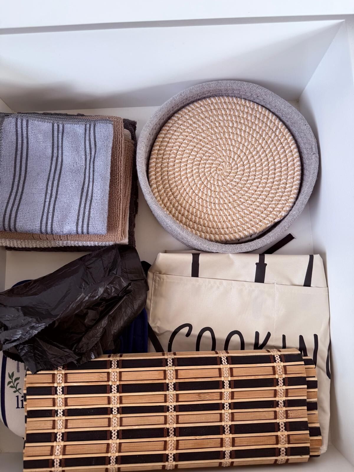 A storage box containing various items including a woven basket, a striped fabric, a black plastic bag, a beige bag with 'COOKIN' text, and a woven tray.