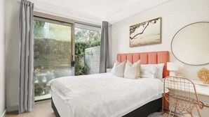 A bedroom with a bed, a desk with a chair, a painting, and a view through a glass door.