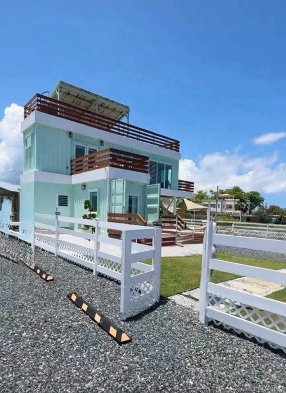 A modern two-story building with a white and light blue color scheme, a balcony on the upper floor, and a white fence in the foreground.