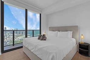Ein modernes Schlafzimmer mit einem großen Bett, einem Nachttisch und Blick auf eine Stadtlandschaft durch ein großes Fenster.