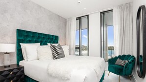 Ett modernt hotellrum med en stor säng, ett nattduksbord, en grön fåtölj och utsikt över havet.