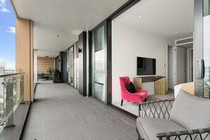 A modern hotel balcony with a glass railing, a seating area with a red chair, a grey sofa, and a flat-screen TV mounted on the wall. - Wynyard Quarter Executive Apt – 3BR Waterfront Living (Auckland City)