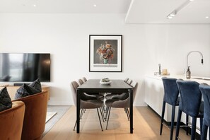 A modern living room with a flat-screen TV, a dining area with a dark wooden table and chairs, and a kitchen with a white countertop and bar stools. - Wynyard Quarter Executive Apt – 3BR Waterfront Living (Auckland City)
