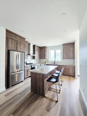 A modern kitchen with wooden cabinets, stainless steel appliances, and a central island with bar stools. - Bright & Homey 3br New Home in NW Calgary (Calgary)
