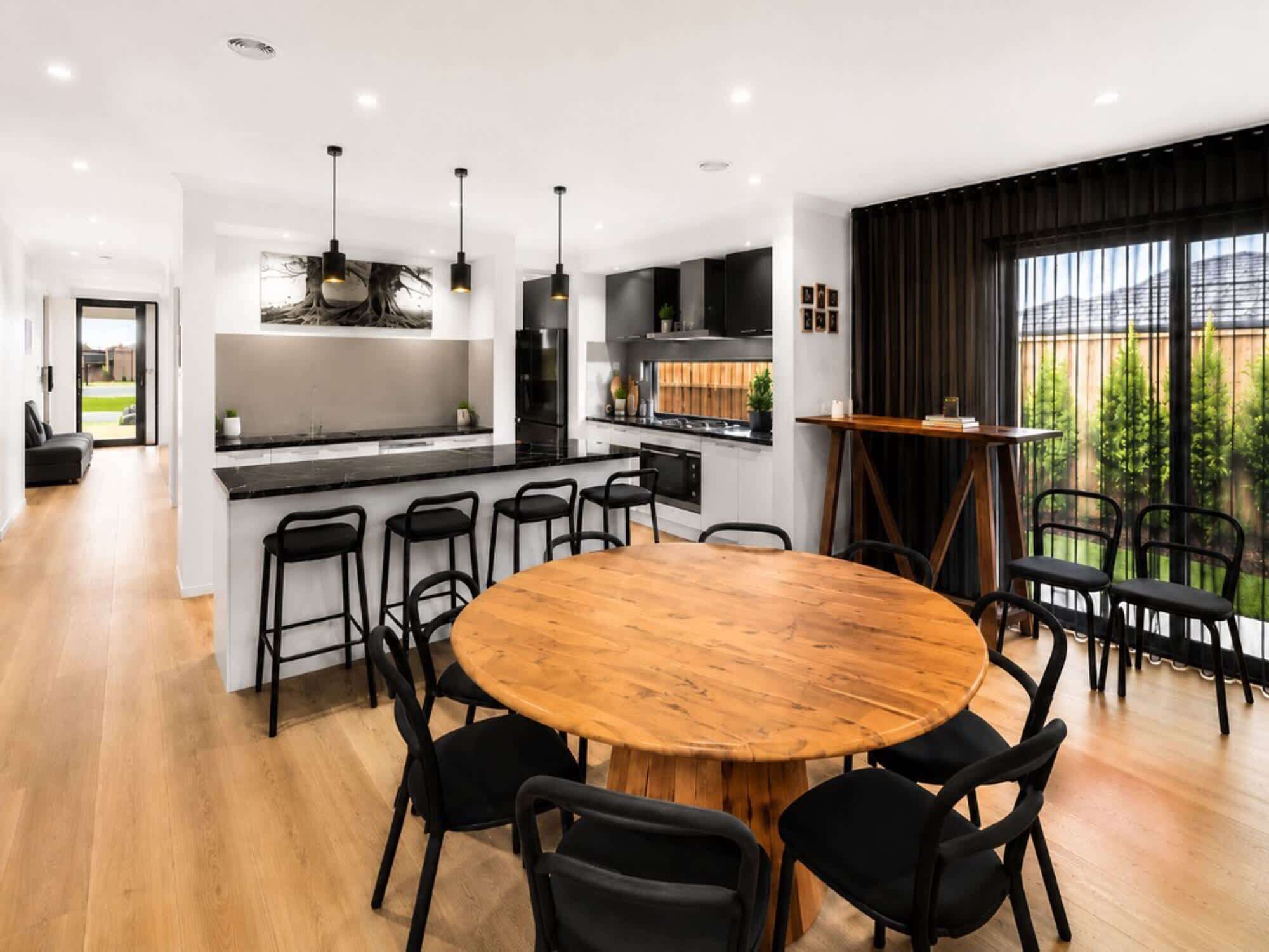 A modern kitchen with a central island, black bar stools, a round wooden dining table, and a view of a garden through large windows.
