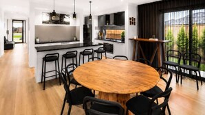 Una cuina moderna amb una illa central, tamborets negres, taula rodona de fusta i vistes a un jardí a través de grans finestres.