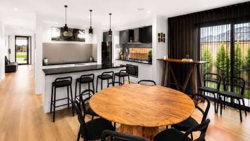 A modern kitchen with a central island, black bar stools, a round wooden dining table, and a view of a garden through large windows.