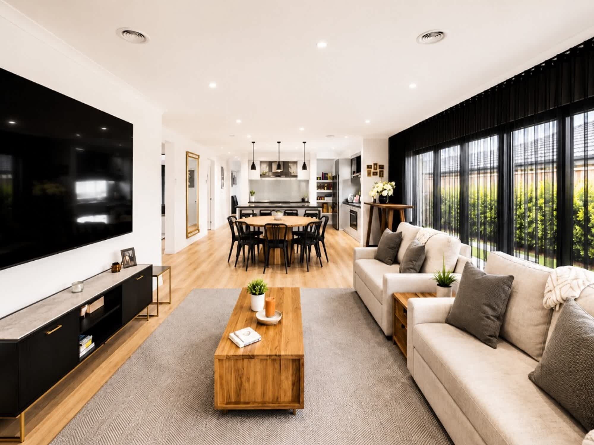 A modern living room with a large TV, a wooden coffee table, a sofa, and a dining area with chairs and a kitchen in the background.