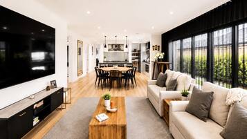 A modern living room with a large TV, a wooden coffee table, a sofa, and a dining area with chairs and a kitchen in the background.