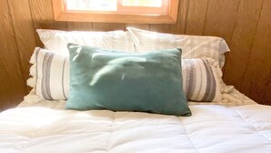 A bed with white bedding and a teal pillow, wooden paneling on the walls, and a window above the bed.