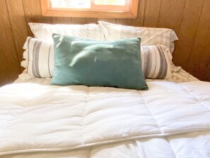 Ein Bett mit weißer Bettwäsche und einem türkisfarbenen Kopfkissen, Holzpaneele an den Wänden und ein Fenster über dem Bett.