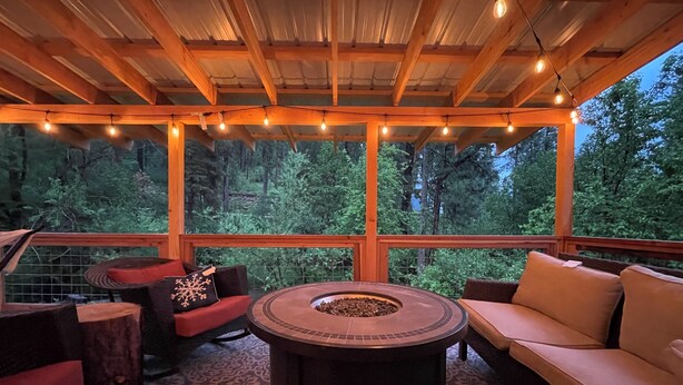 A covered outdoor seating area with a fire pit, wooden furniture, and a view of the forest.