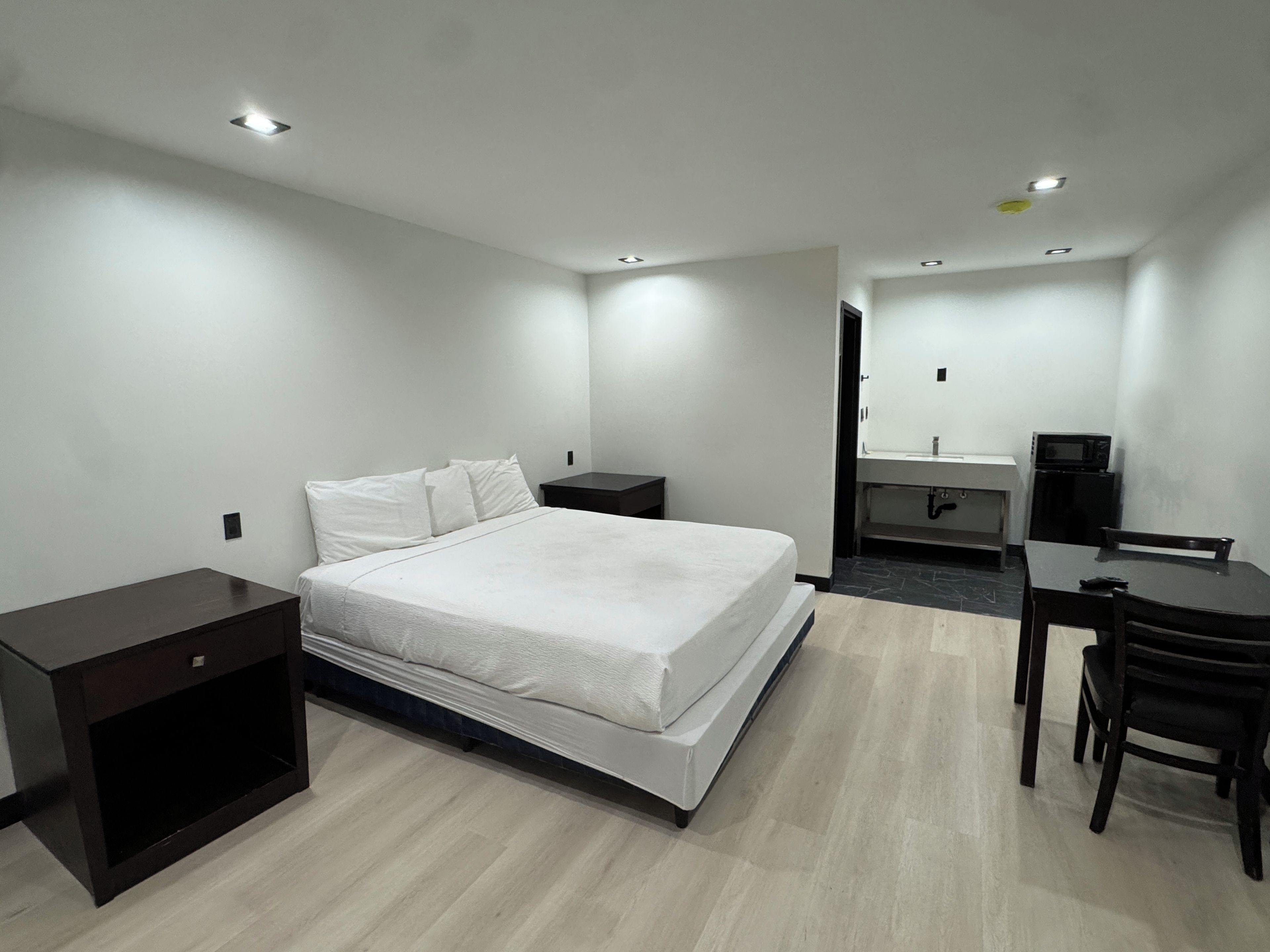 A modern hotel room with a large bed, a desk, a chair, a small kitchenette, and a bathroom.