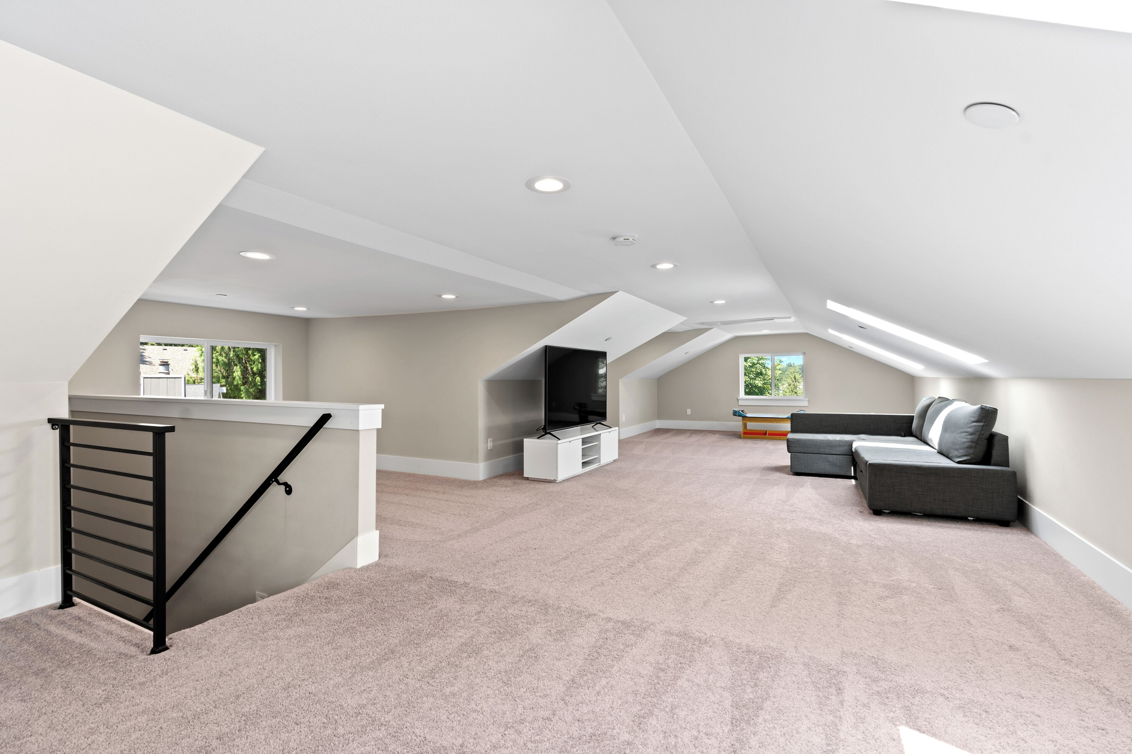 A spacious attic room with a high ceiling, a built-in entertainment unit, a grey sectional sofa, and a staircase with a black handrail.