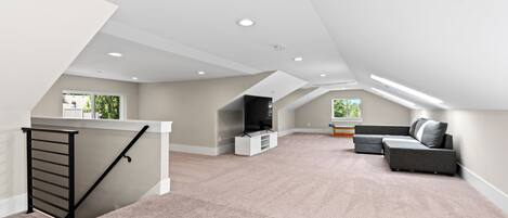 A spacious attic room with a high ceiling, a built-in entertainment unit, a grey sectional sofa, and a staircase with a black handrail.