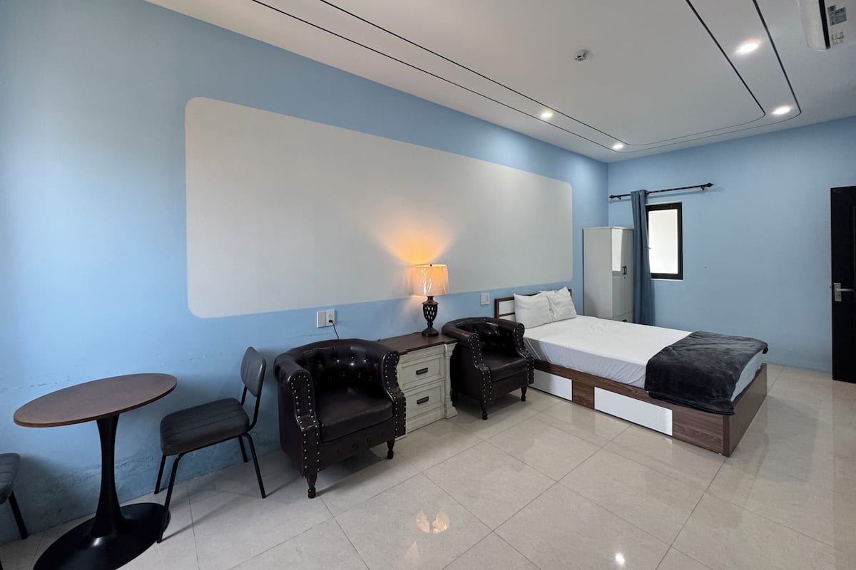 Habitación de hotel con cama, escritorio, silla, mesita, lámpara, armario y ventana.