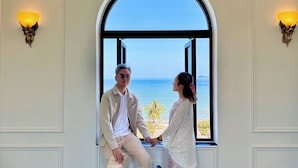 A couple holding hands in a room with a large arched window overlooking the sea, marble flooring, and wall-mounted sconces.