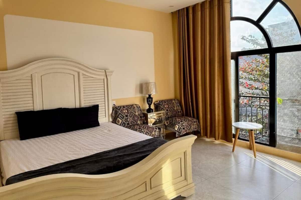 Un dormitorio con una cama, una mesita de noche, una silla, una pequeña mesa y un ventanal grande.