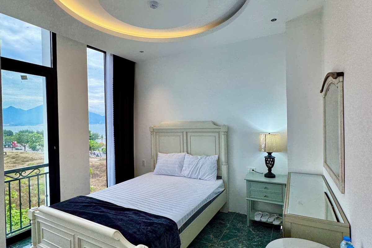 Habitación de hotel con cama, una mesa redonda, una silla, una mesita de noche, una lámpara, un espejo y un balcón con vistas.