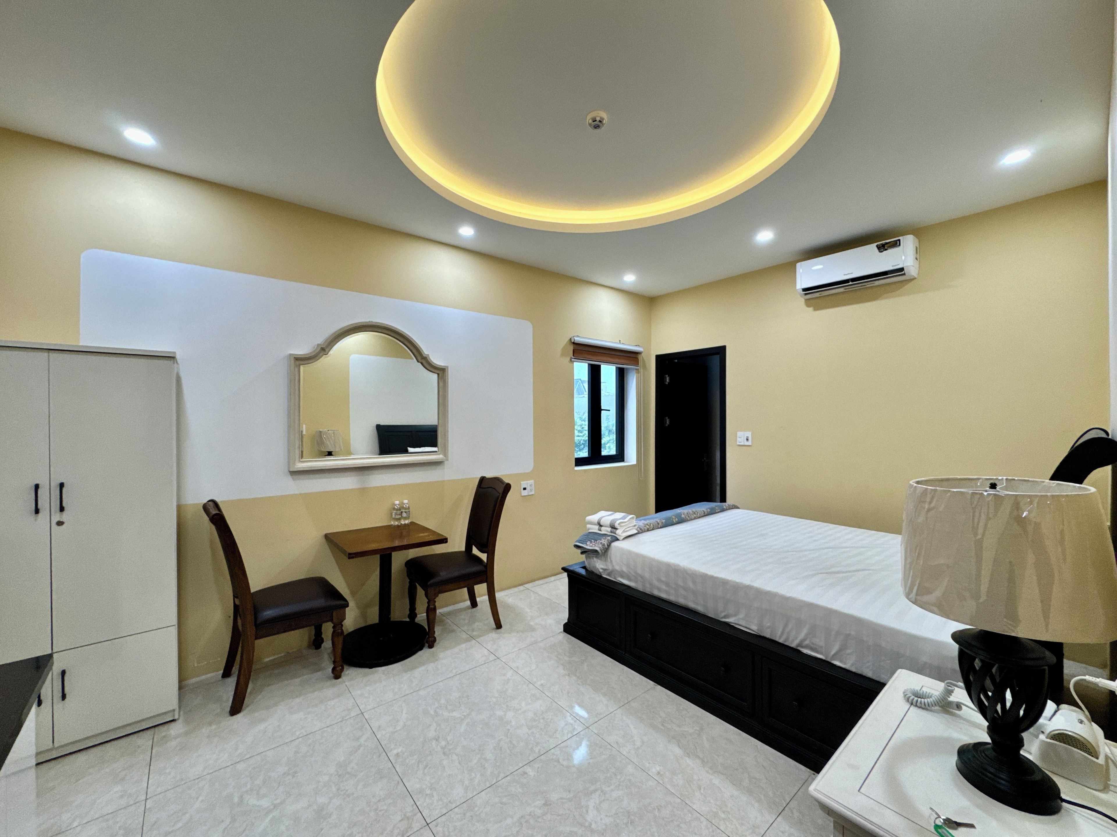 A hotel room with a bed, a desk, a chair, a wardrobe, a mirror, a window, and an air conditioning unit.
