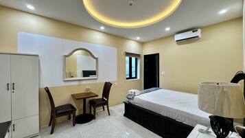 A hotel room with a bed, a desk, a chair, a wardrobe, a mirror, a window, and an air conditioning unit.