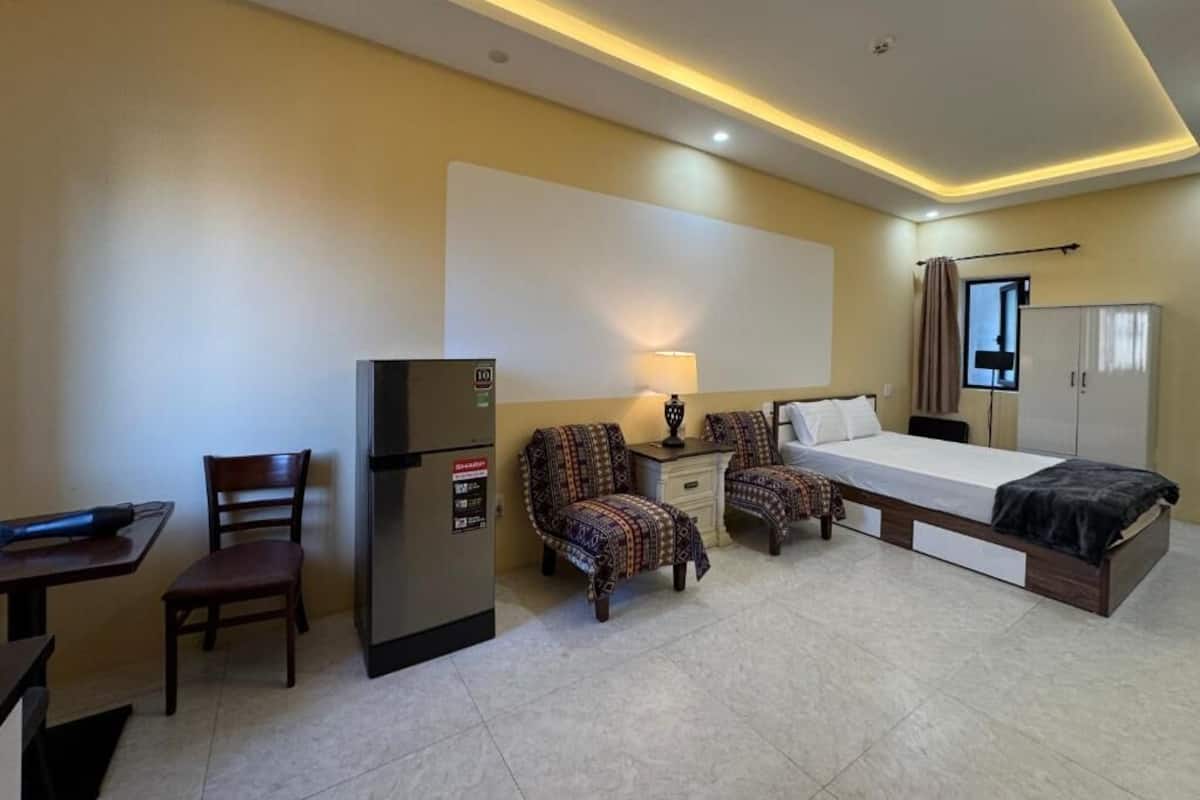 Habitación de hotel con cama, escritorio con silla, refrigerador y mesita de noche con lámpara.