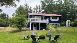 Une maison à deux étages avec un toit rouge, un balcon et une cour verdoyante où se trouvent quatre chaises Adirondack.